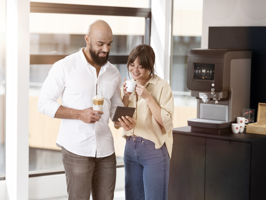 To personer ved kaffemaskin ser på nettbrett i moderne pauseområde.
