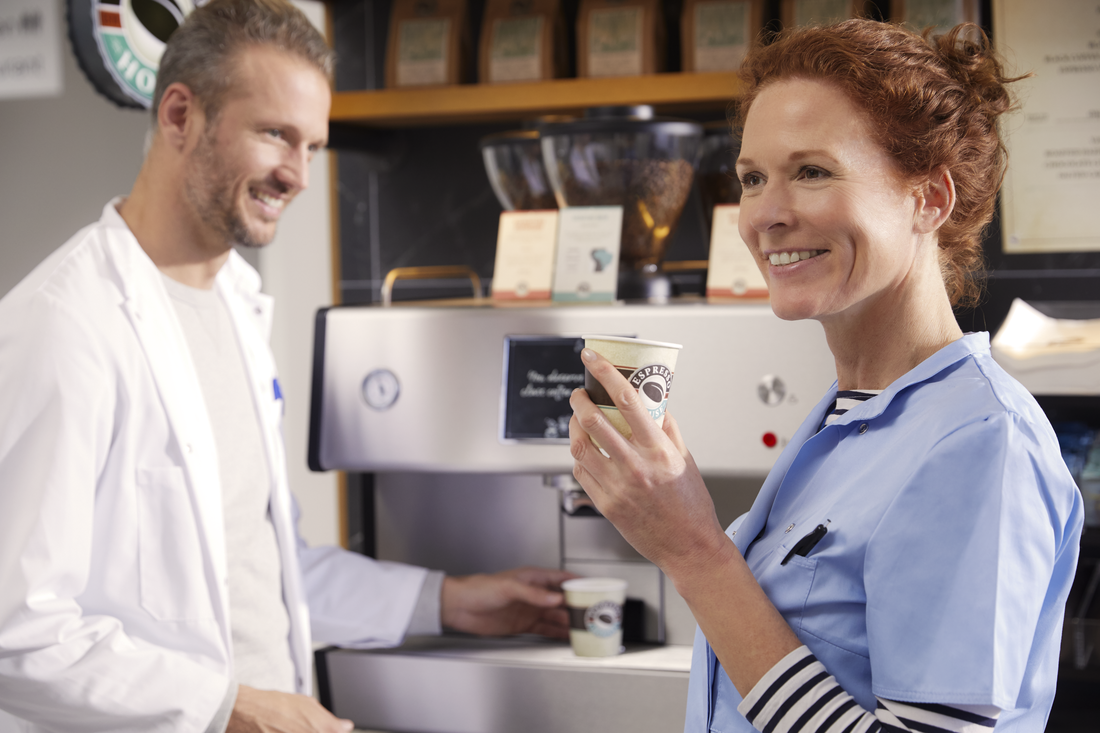 To personer ved en kaffemaskin, én i hvit frakk og én i blått arbeidstøy. Kaffeposer på hylle over maskinen.